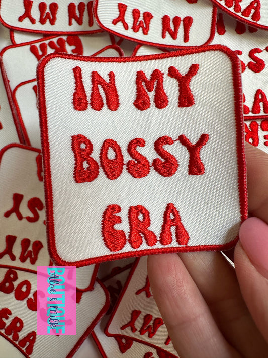A hand holds a square embroidered patch with red lettering that reads 'IN MY BOSSY ERA' on a cream white background with red border. Multiple similar patches are scattered in the background.