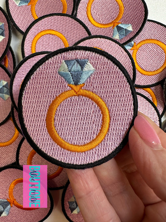 A close-up of a hand holding a round embroidered patch featuring a pink background, a gold ring, and a diamond, with other matching patches visible behind it.